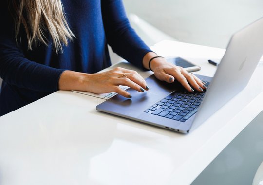 Person arbeitet an einem Laptop auf einem hellen Schreibtisch, daneben liegen Smartphone und Notizzettel.
