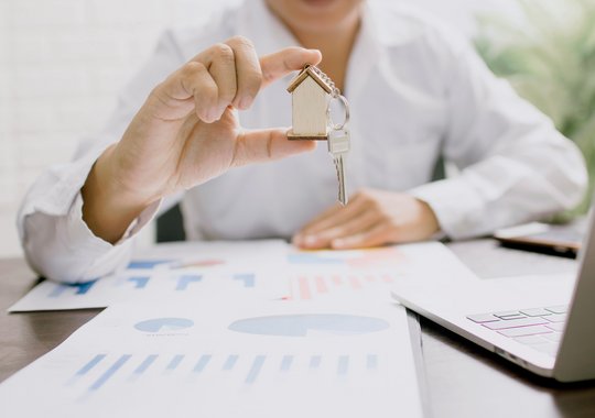 Hand hält Schlüssel mit Hausanhänger über Dokumenten und Laptop, Symbol für Immobilienkauf 