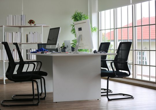 Modernes Büro mit großem Schreibtisch, vier schwarzen Bürostühlen und zwei Monitoren vor bodentiefen Fenstern mit Blick auf ein Haus mit rotem Ziegeldach.