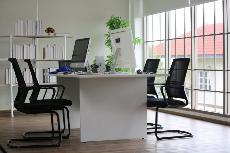 Helles Büro mit modernem Arbeitsplatz Modernes Büro mit großem Schreibtisch, vier schwarzen Bürostühlen und zwei Monitoren vor bodentiefen Fenstern mit Blick auf ein Haus mit rotem Ziegeldach.