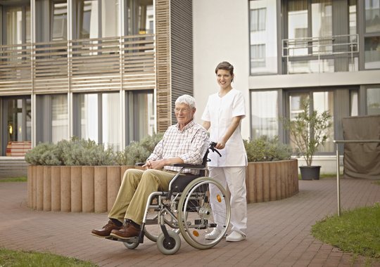 Altenpflegerin mit Rollstuhl vor einer Immobilie 