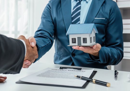 Immobilienberater im Anzug überreicht ein Hausmodell und besiegelt den Vertragsabschluss mit Handschlag über einem unterschriebenen Immobilienvertrag.