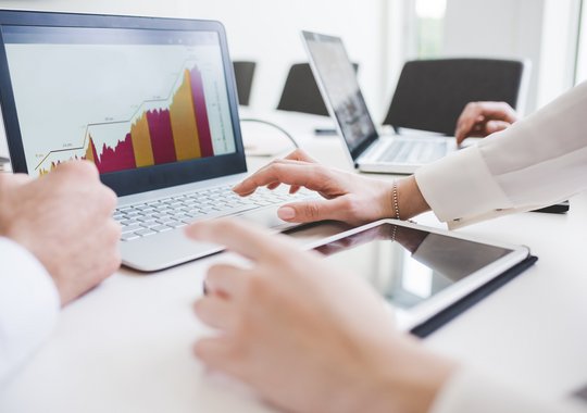 Personen analysieren auf einem Laptop ein farbiges Balken- und Liniendiagramm mit Wachstumsentwicklung; im Vordergrund ein Tablet, im Hintergrund weitere Laptops.