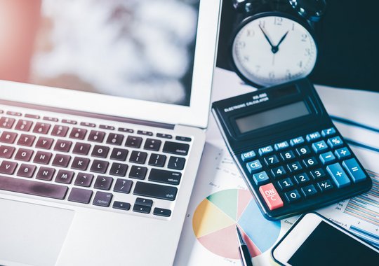 Schreibtisch mit Laptop, Taschenrechner, Uhr und Geschäftsunterlagen mit Diagrammen und Kreisdiagramm, daneben ein Smartphone und ein Kugelschreiber.