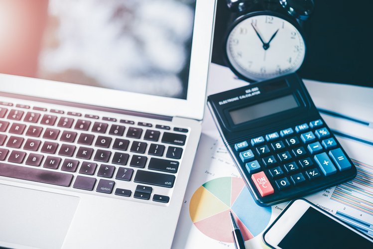 Büroarbeitsplatz_mit_Technik_und_Unterlagen Schreibtisch mit Laptop, Taschenrechner, Uhr und Geschäftsunterlagen mit Diagrammen und Kreisdiagramm, daneben ein Smartphone und ein Kugelschreiber.